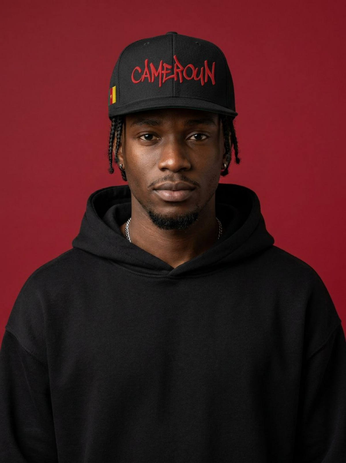 Homme portant une casquette snapback noir avec Cameroun brodé en rouge et le drapeau camerounais, hoodie noir, fond rouge.